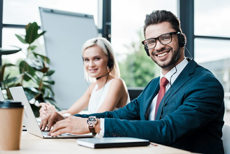 selective focus of cheerful man in headset typing on laptop while sitting near blonde colleague